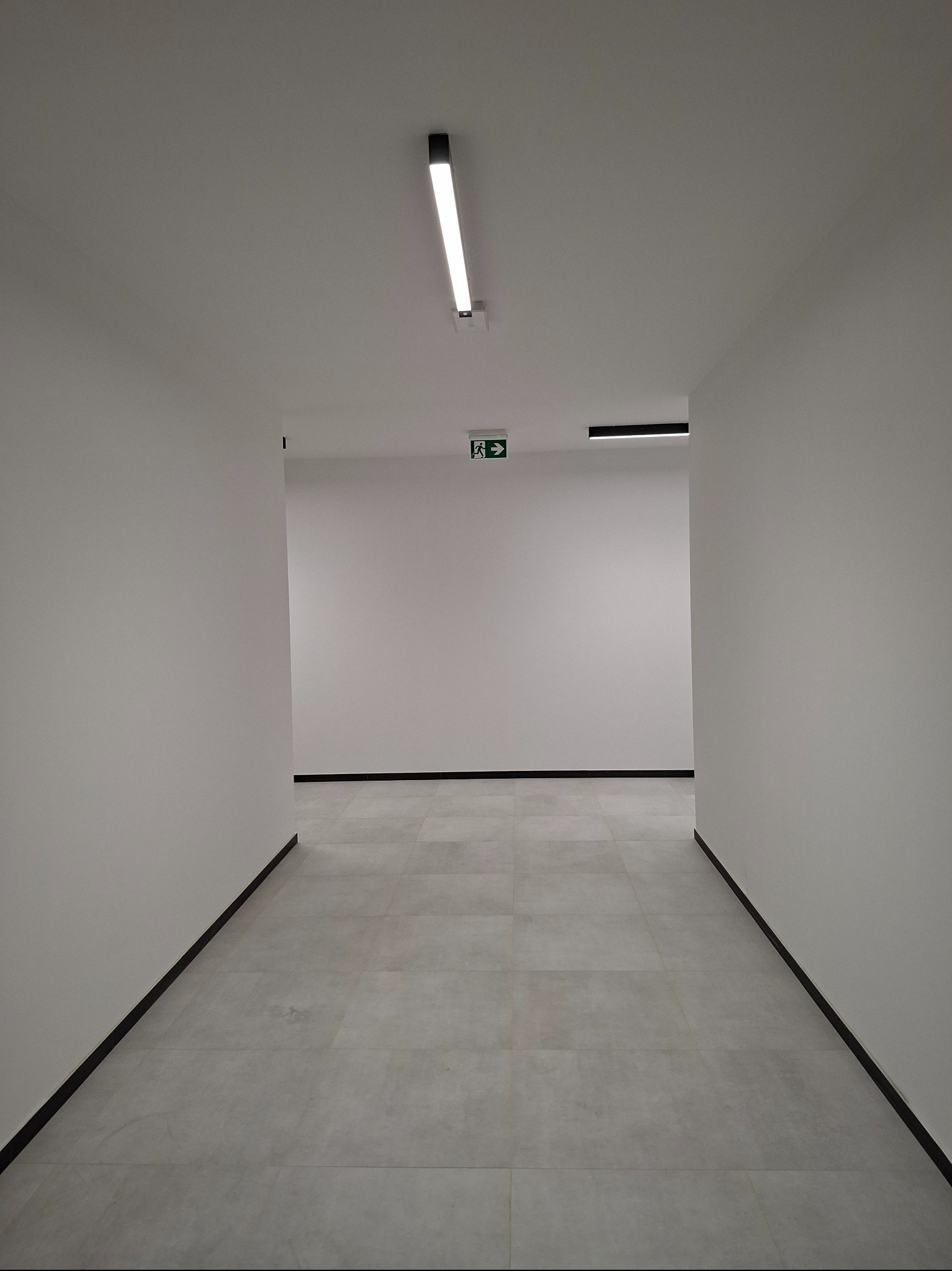 Biały korytarz z szarą podłogą z płytek, oświetlony lampą LED. Widoczny znak ewakuacyjny. Minimalistyczne wykończenie wnętrza, świeżo po remoncie.