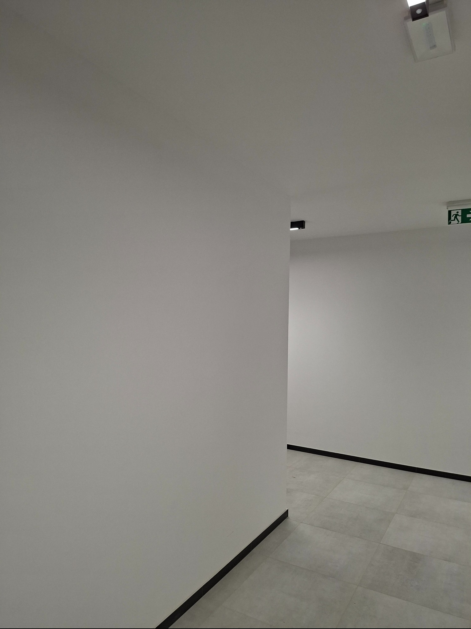 Białe ściany korytarza z szarą podłogą z płytek. Widoczny znak ewakuacyjny i czarne listwy przypodłogowe. Minimalistyczne oświetlenie sufitowe.