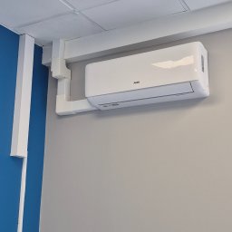 NERIN - Klimatyzacja - Biały klimatyzator ścienny AUX zamontowany na szarej ścianie z widocznymi białymi maskownicami instalacyjnymi biegnącymi do sufitu podwieszanego. Fragment niebieskiej ściany po lewej.