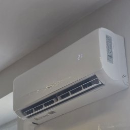 NERIN - Klimatyzacja - Biała, nowoczesna jednostka klimatyzacyjna zamontowana wysoko na jasnej ścianie, z cyfrowym wyświetlaczem '21' i otwartymi żaluzjami nawiewu, ukazująca gotowość do pracy.