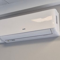NERIN - Klimatyzacja - Biała jednostka wewnętrzna klimatyzatora ściennego AUX, zamontowana na jasnoszarej ścianie pod sufitem, z widocznym białym korytem instalacyjnym.