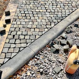 Alan Urban - Fragment układania kostki granitowej: szara kostka, betonowy obrzeżnik, podsypka i kanister. Widoczne postępy prac brukarskich.