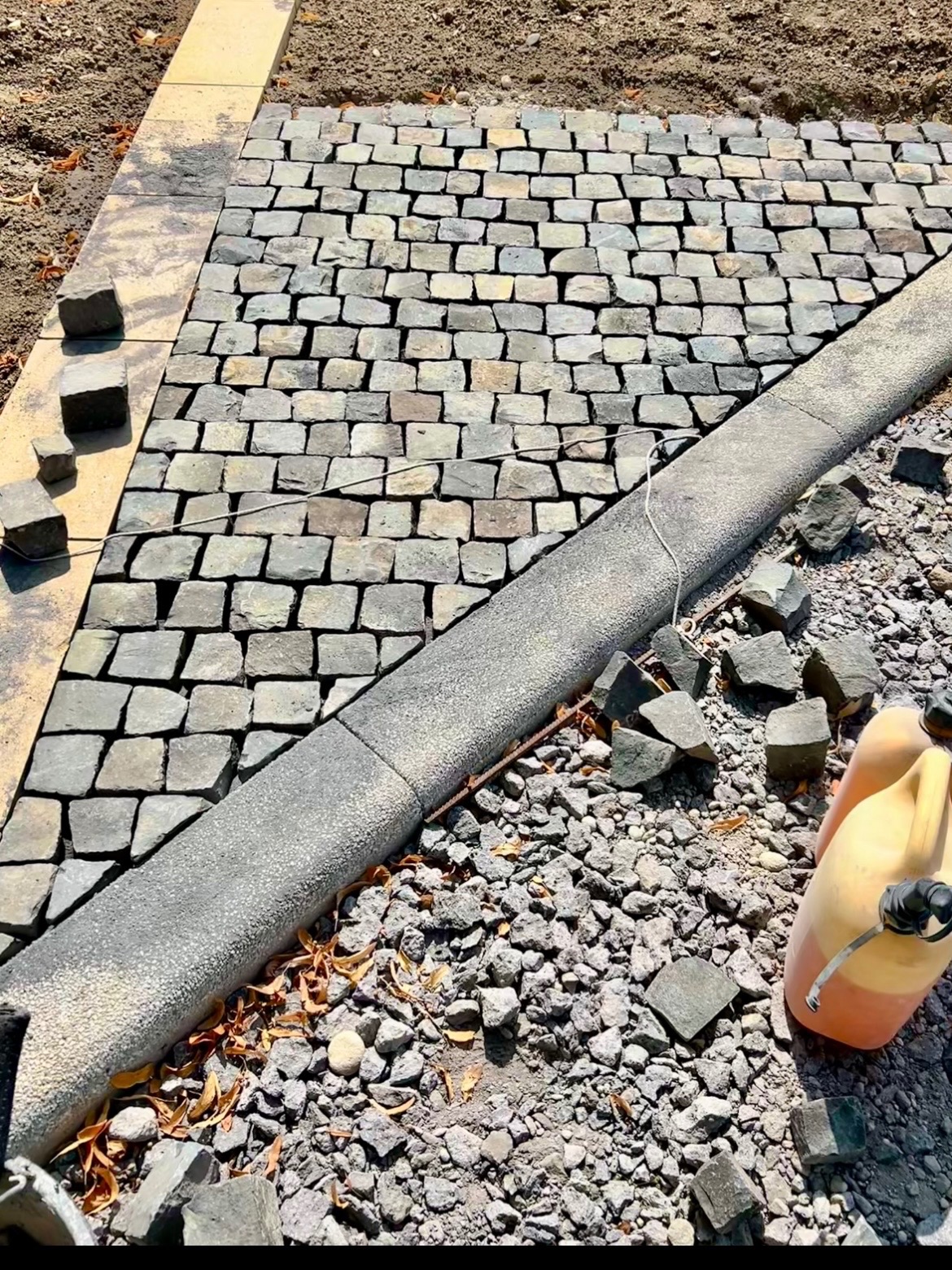 Fragment układania kostki granitowej: szara kostka, betonowy obrzeżnik, podsypka i kanister. Widoczne postępy prac brukarskich.