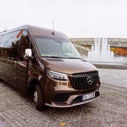 TRANS-TAXI PRZEWÓZ OSÓB I TOWARU PIOTR GRYM - Brązowy, luksusowy bus Mercedes-Benz na tle fontanny i nowoczesnej architektury. Widok z boku, podkreślający elegancki design i przestronność pojazdu.
