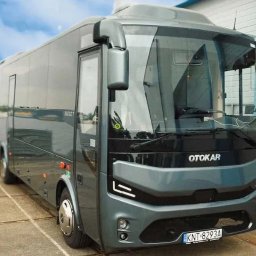 TRANS-TAXI PRZEWÓZ OSÓB I TOWARU PIOTR GRYM - Szary bus Otokar Navigo T z widoczną tablicą rejestracyjną KNT 8293A, zaparkowany na zewnątrz budynku z niebieskimi akcentami. Nowoczesny design pojazdu.