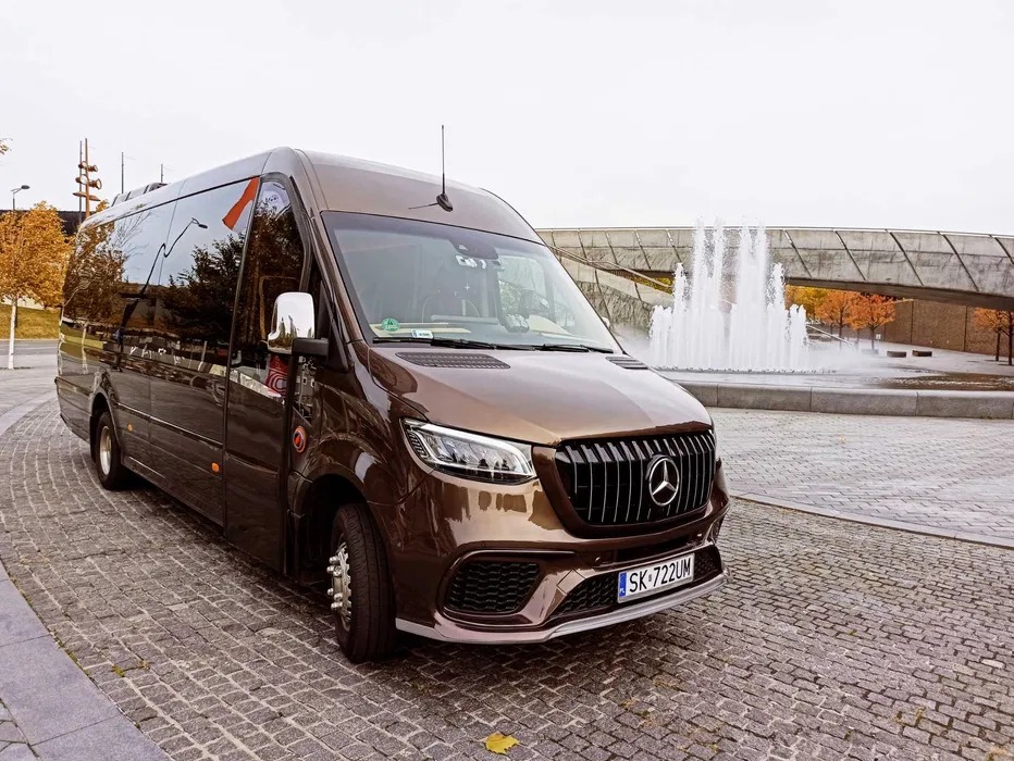 Brązowy, luksusowy bus Mercedes-Benz na tle fontanny i nowoczesnej architektury. Widok z boku, podkreślający elegancki design i przestronność pojazdu.