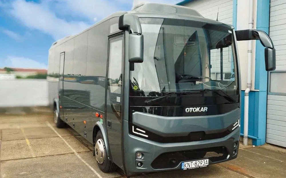 Szary bus Otokar Navigo T z widoczną tablicą rejestracyjną KNT 8293A, zaparkowany na zewnątrz budynku z niebieskimi akcentami. Nowoczesny design pojazdu.