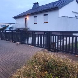 JK STAHL BAU - Ciemne, pionowe ogrodzenie z bramą wjazdową, widoczne z poziomu chodnika z kostki brukowej, na tle białego domu z szarym dachem i zielenią. Ujęcie z dołu.