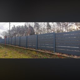 JK STAHL BAU - Długie, poziome ogrodzenie z szarych paneli, z widocznymi przerwami między deskami, biegnące wzdłuż zielonego terenu i drzew. Słupki ogrodzenia w tym samym kolorze.