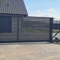 JK STAHL BAU - Nowoczesna, przesuwna brama wjazdowa z poziomych profili, w kolorze grafitowym, na posesji w Namyślinie. Widoczny automat do bramy i fragment ogrodzenia panelowego.