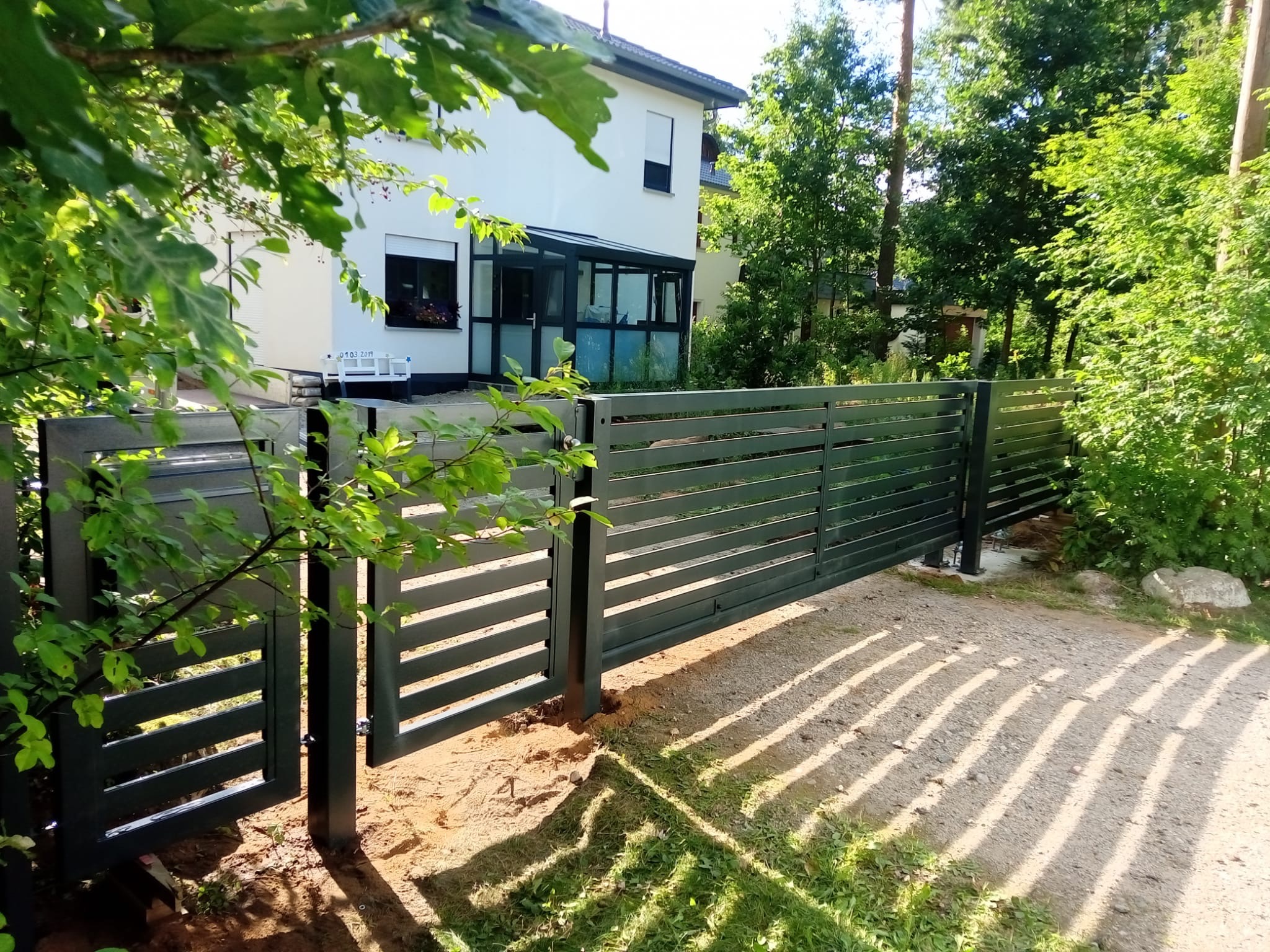 Nowoczesna, pozioma brama wjazdowa z metalowymi przęsłami, częściowo otwarta, na tle białego domu i zieleni. Widoczne ślady montażu w ziemi.