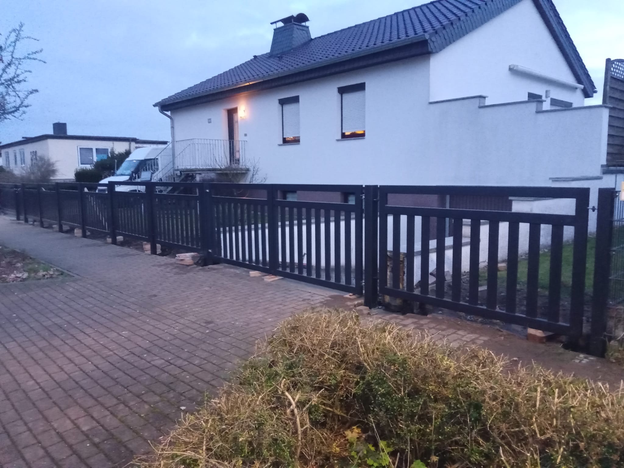 Ciemne, pionowe ogrodzenie z bramą wjazdową, widoczne z poziomu chodnika z kostki brukowej, na tle białego domu z szarym dachem i zielenią. Ujęcie z dołu.