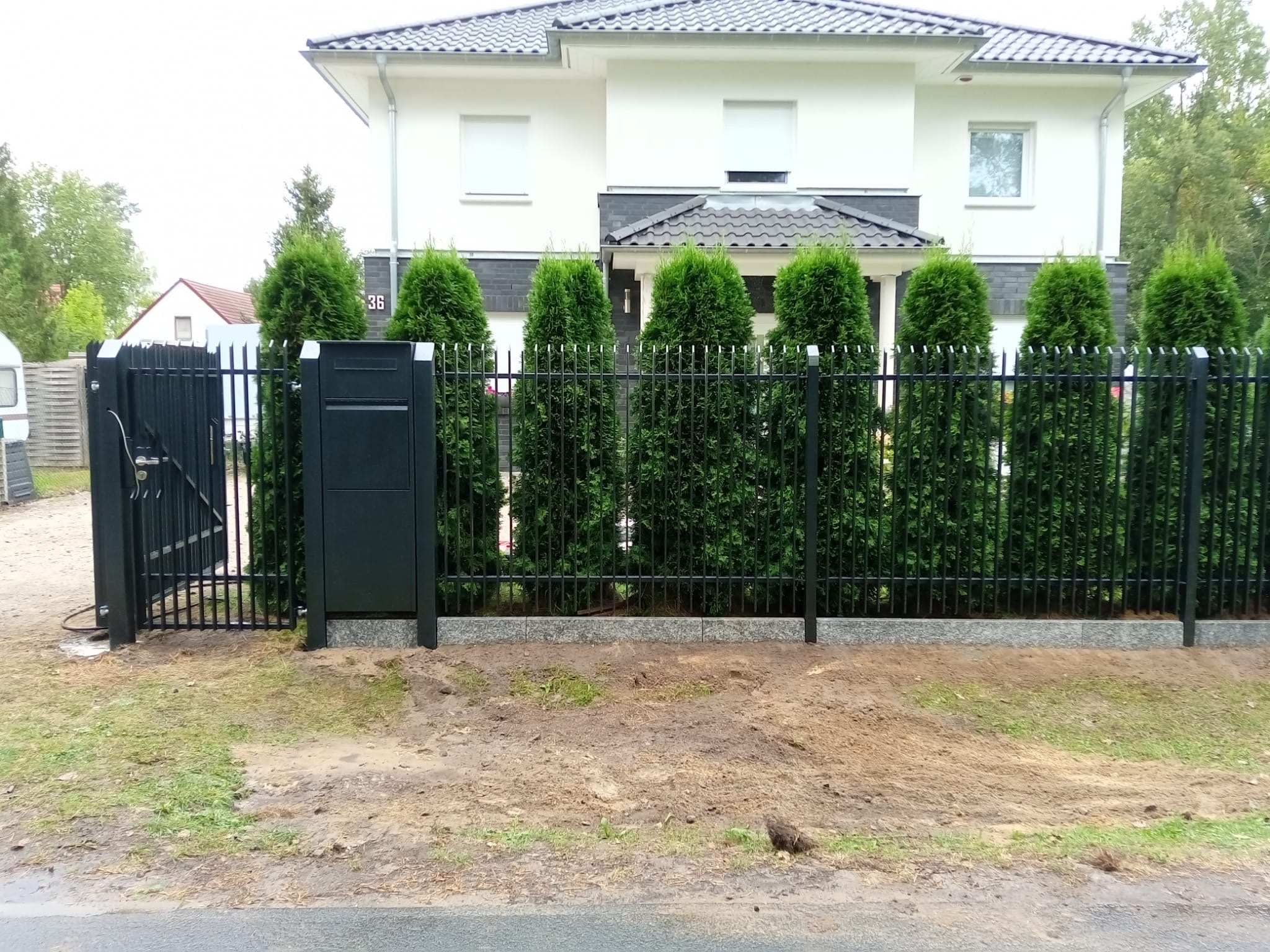 Nowoczesne, czarne ogrodzenie metalowe z furtką i skrzynką na listy, na tle domu z numerem 36 i rzędem tuj. Widok z poziomu gruntu.