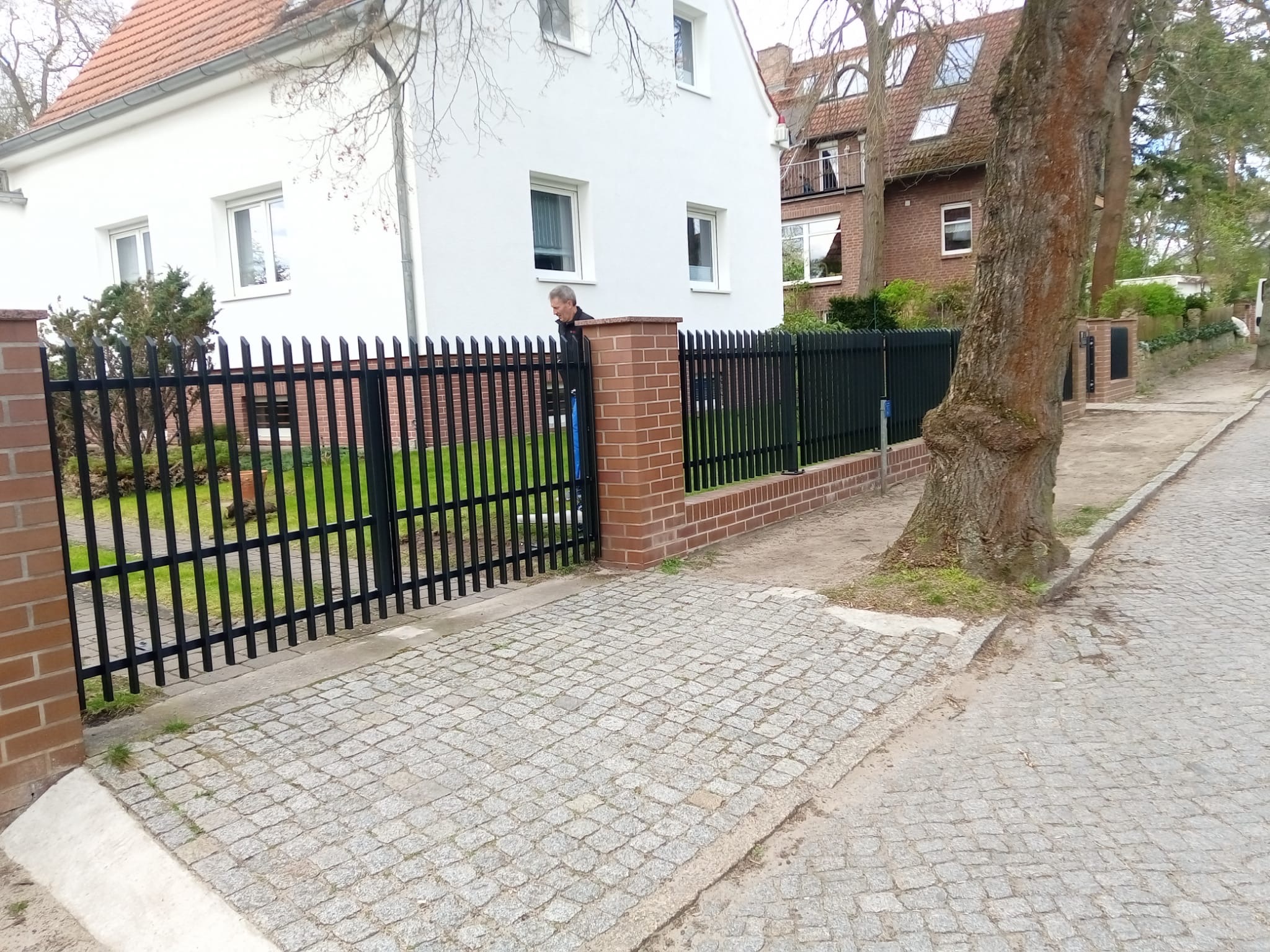 Czarne, metalowe ogrodzenie z ceglanymi słupkami, oddzielające posesję od chodnika wyłożonego kostką. Widoczny fragment domu z białymi ścianami i czerwonym dachem.