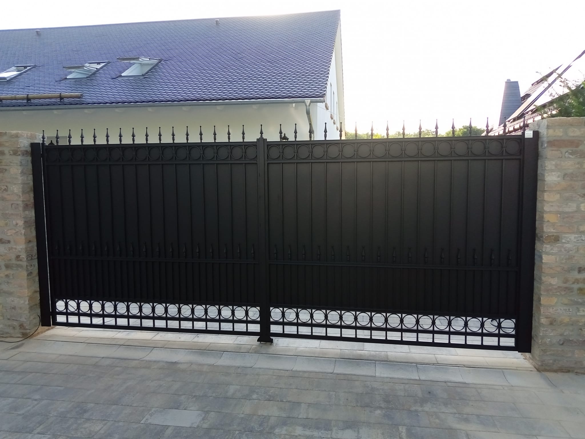 Czarna, metalowa brama wjazdowa z dekoracyjnymi elementami, osadzona między murowanymi słupkami. Widoczny fragment domu z ciemnym dachem i jasną elewacją.