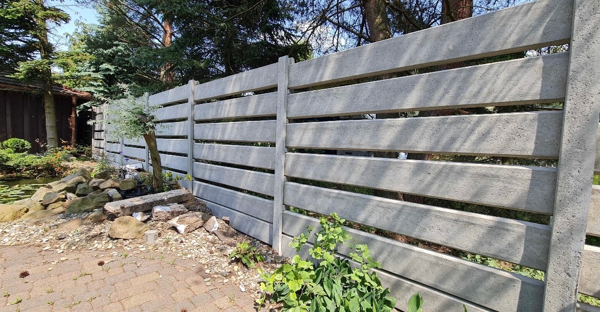 Nowoczesne ogrodzenie betonowe z poziomymi panelami w Namyślinie, otoczone zielenią. Widoczne elementy ogrodu i drzewa w tle. Minimalistyczny design.