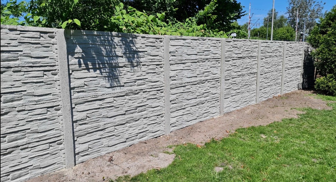 Szary, betonowy płot o fakturze kamienia, z widocznymi słupkami, oddzielający trawnik od zieleni. Widok z bliska, podkreślający strukturę materiału.