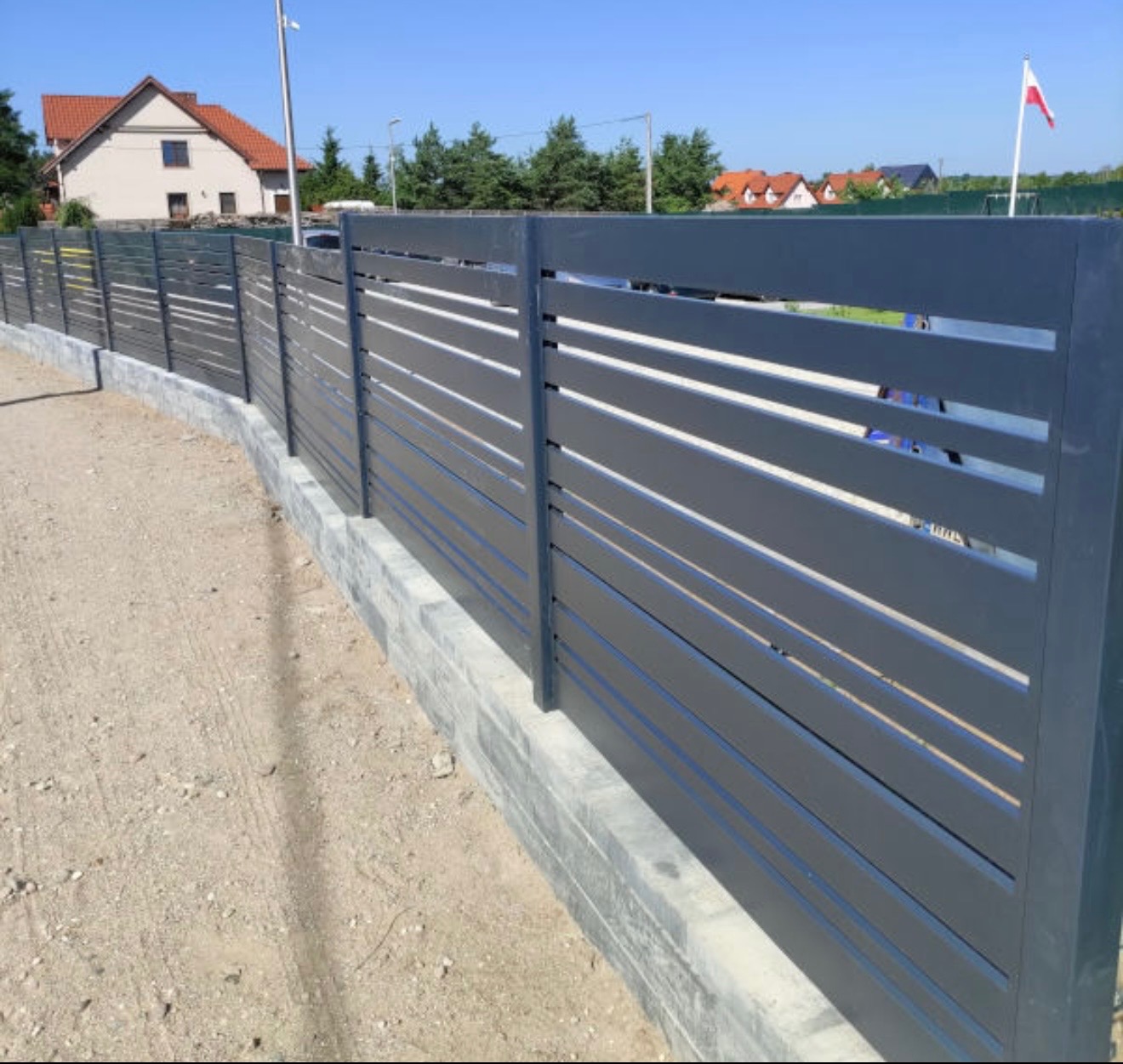 Nowoczesne, poziome ogrodzenie metalowe w kolorze grafitowym, osadzone na betonowej podmurówce. W tle dom z czerwonym dachem i maszt z flagą na tle błękitnego nieba.
