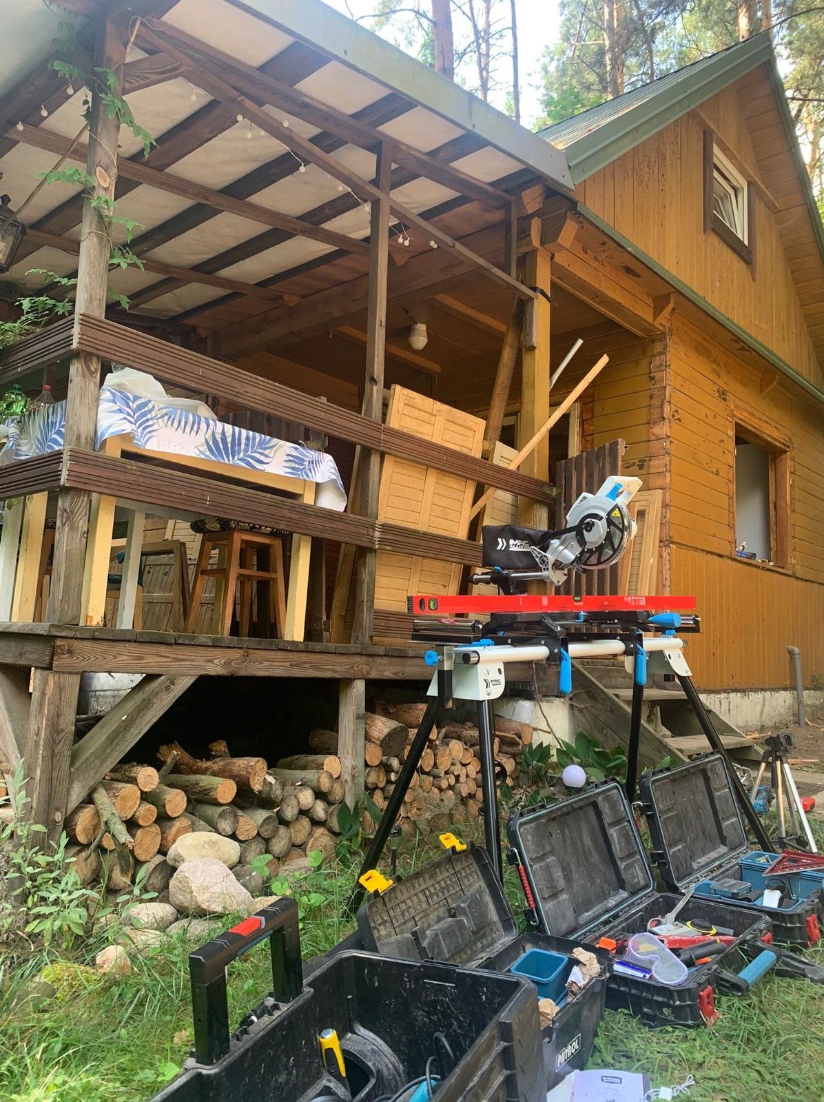 Drewniany dom z werandą w trakcie budowy/remontu. Widoczne narzędzia: piła, poziomica, skrzynki narzędziowe. Na werandzie leżą elementy konstrukcyjne.
