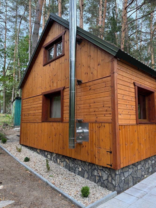 Drewniany domek letniskowy z kominem na tle drzew. Elewacja z desek w ciepłym odcieniu brązu, kamienna podmurówka i okna z drewnianymi ramami. Otoczenie leśne.