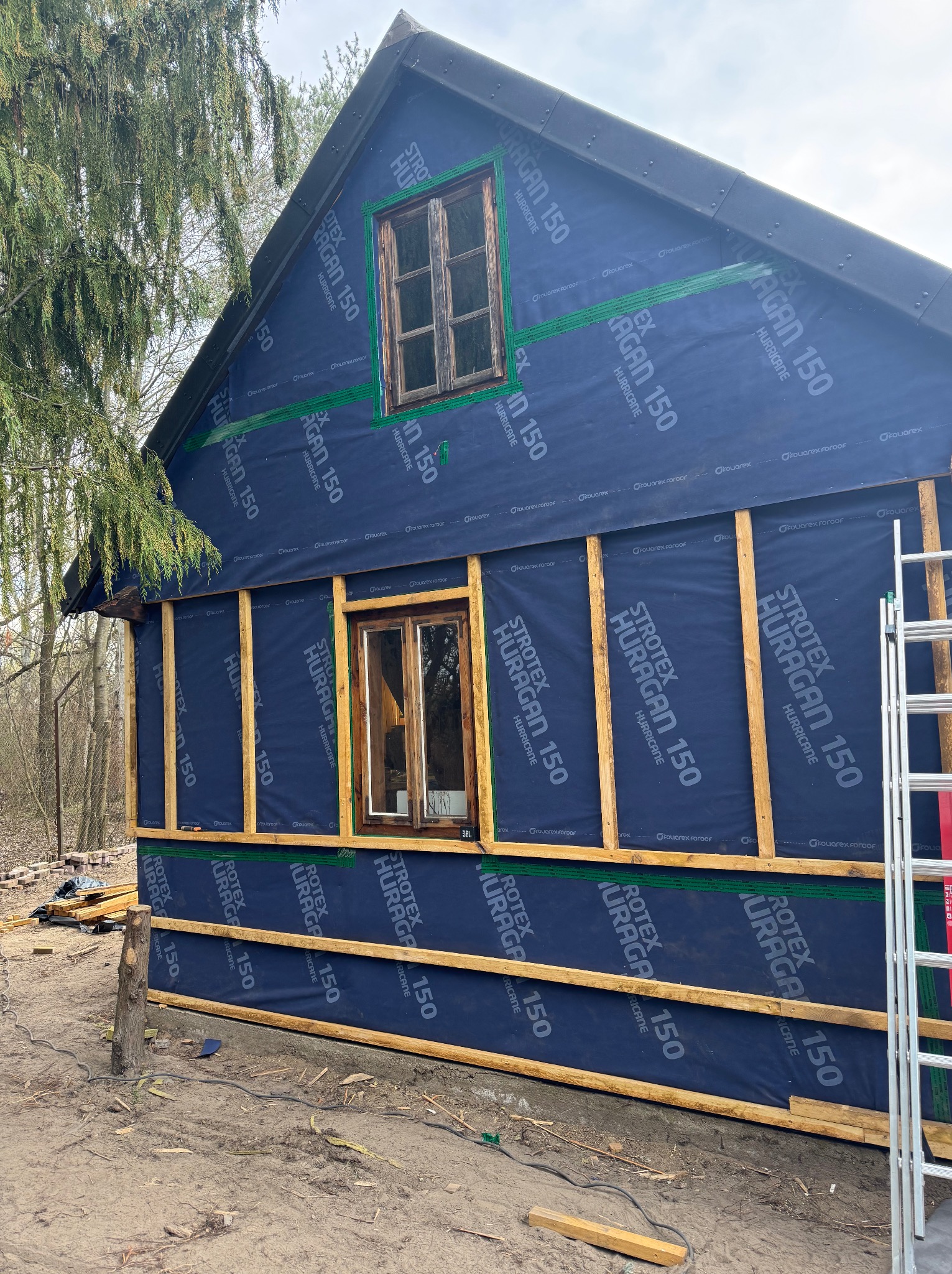 Budowa domu szkieletowego: montaż folii wiatroizolacyjnej Stroetex Huragan 150 na drewnianej konstrukcji. Widoczne okna i drabina. Prace w toku.