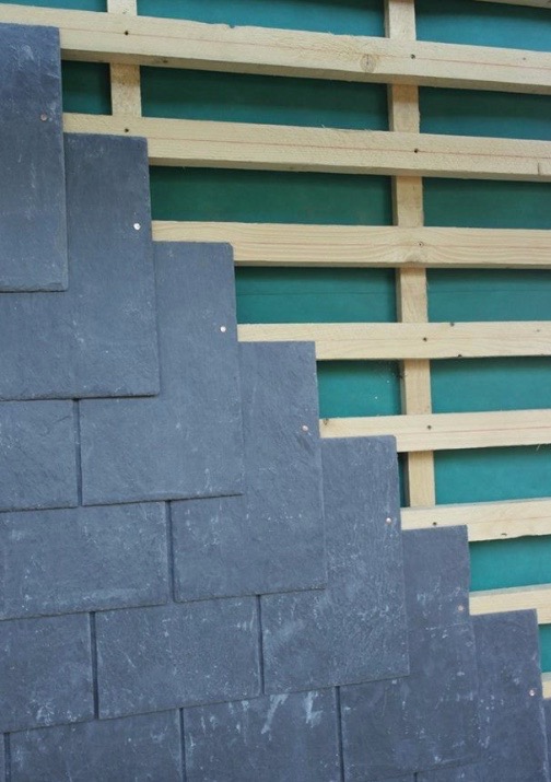 Układanie ciemnych, prostokątnych płyt na drewnianej konstrukcji z zielonym podkładem. Widoczne gwoździe mocujące. Detal dachu w trakcie budowy.