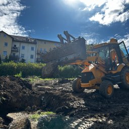 Roboty ziemne Pagacz - Żółta koparko-ładowarka JCB z pełną łyżką ziemi na placu budowy, w tle budynek mieszkalny i błękitne niebo z chmurami. Widoczne ślady opon na błotnistym terenie.