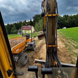 Roboty ziemne Pagacz - Żółta koparka i wywrotka Terex w trakcie prac ziemnych na tle zieleni i pochmurnego nieba, widok z kabiny operatora. Prace ziemne w toku.