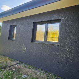 Was&Bud - Elewacja domu jednorodzinnego z ciemnoszarym tynkiem strukturalnym, widoczne dwa okna z czarnymi ramami i rura spustowa. Na oknie żółta tablica informacyjna.