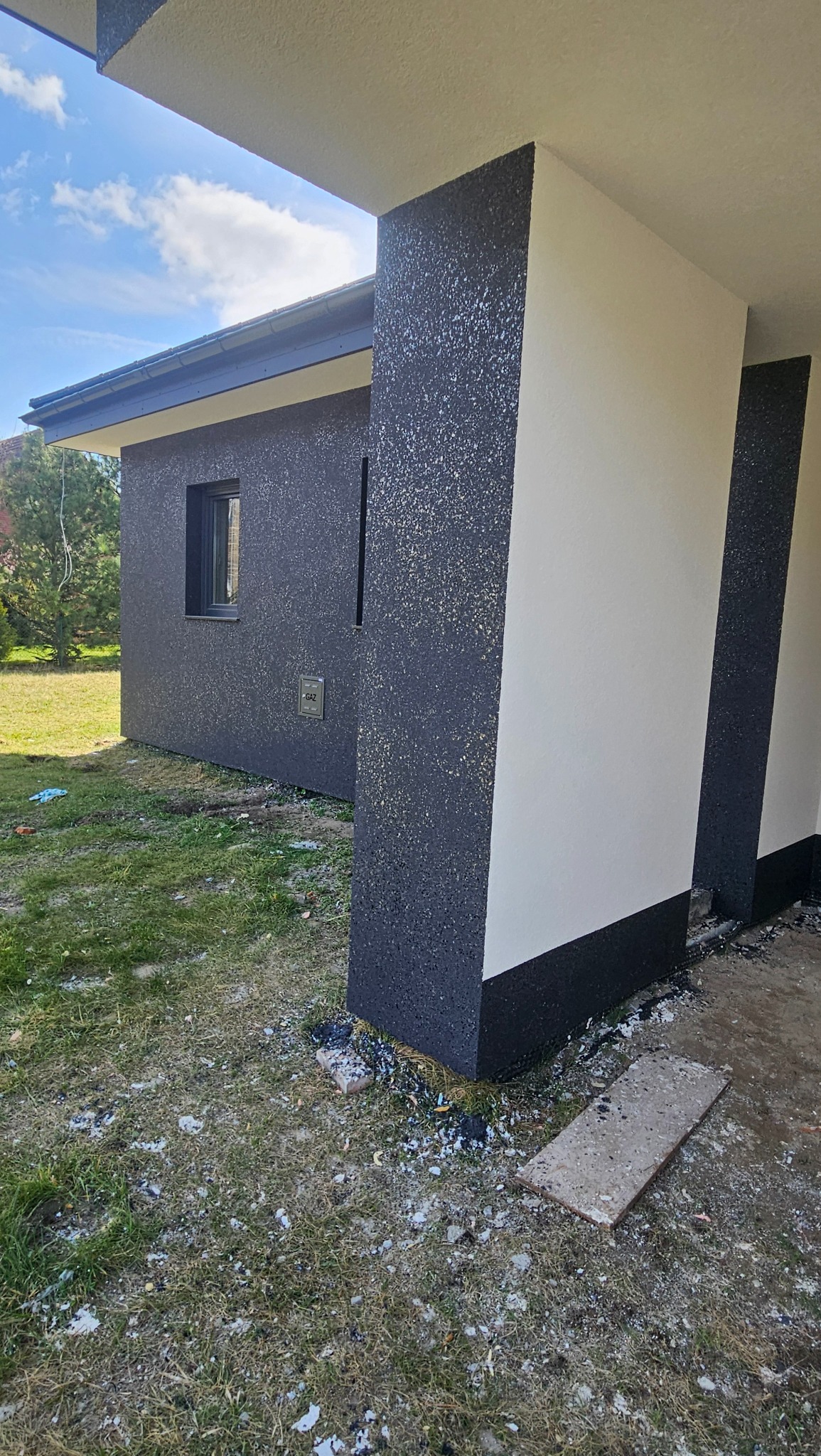 Nowoczesna elewacja domu z kontrastowym tynkiem strukturalnym w odcieniach szarości i bieli. Widoczne detale wykończenia i fragment trawnika.