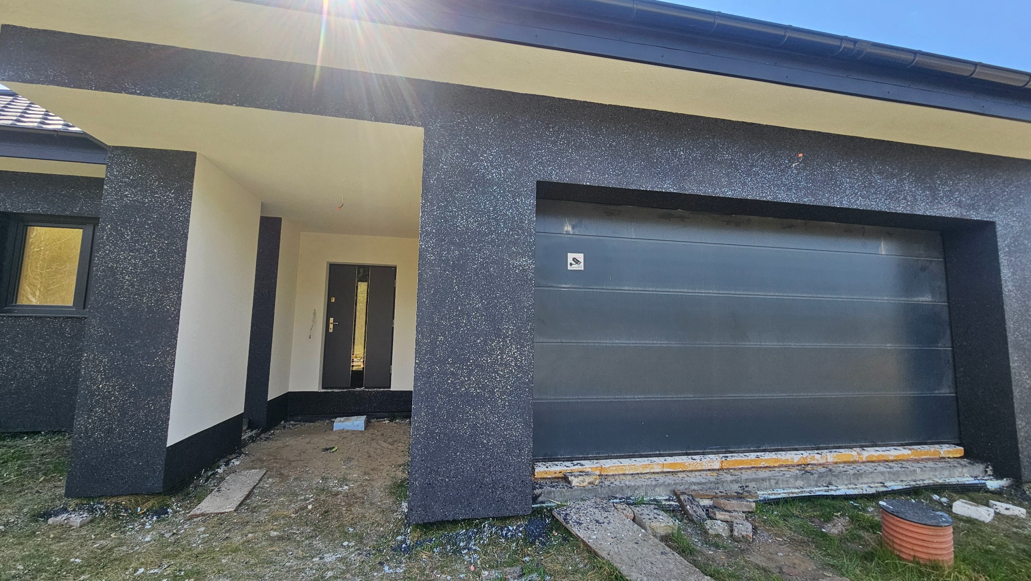 Nowoczesny dom z elewacją w odcieniach szarości i bieli, widoczna brama garażowa i wejście. Prace wykończeniowe w toku, widoczne materiały budowlane.