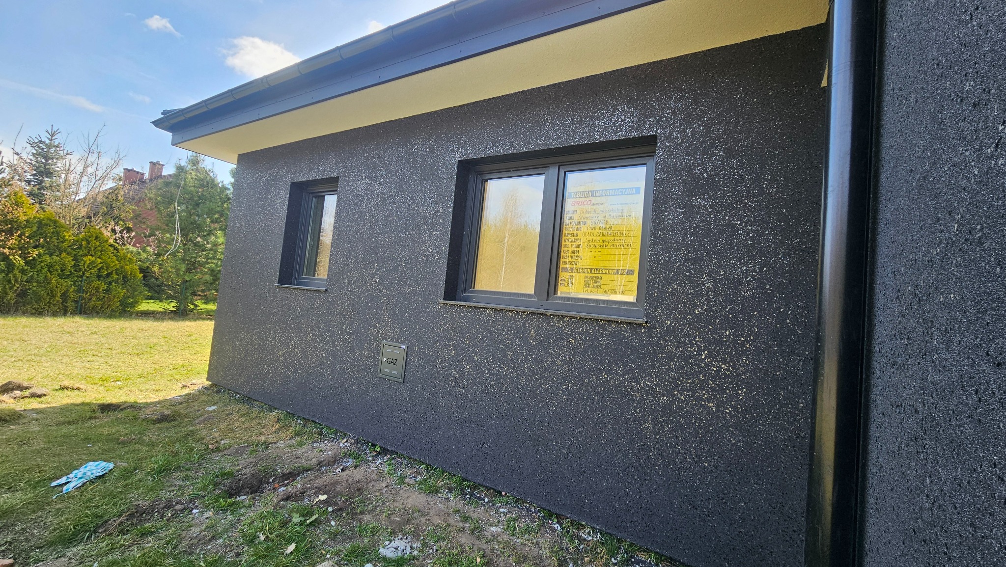 Elewacja domu jednorodzinnego z ciemnoszarym tynkiem strukturalnym, widoczne dwa okna z czarnymi ramami i rura spustowa. Na oknie żółta tablica informacyjna.