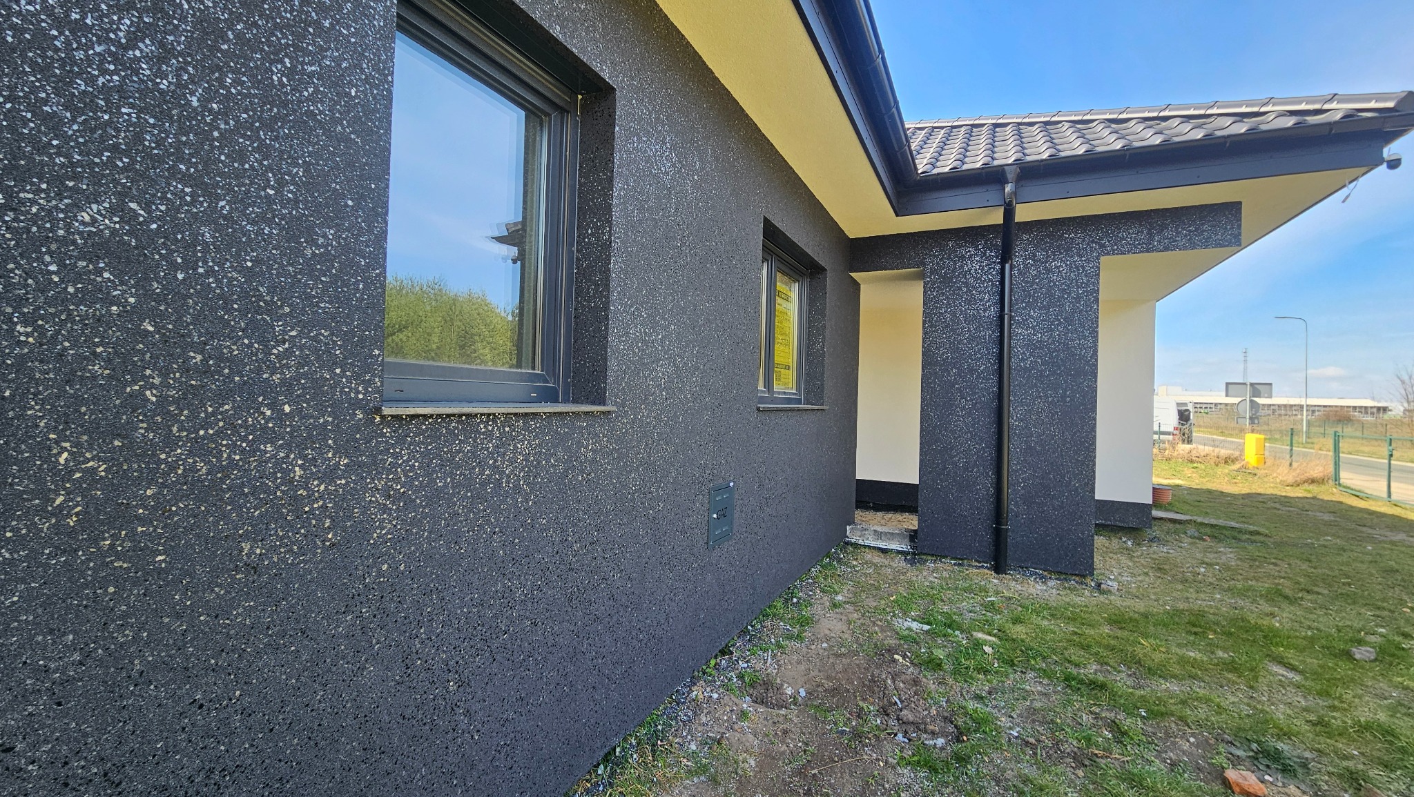 Elewacja domu jednorodzinnego z oknami, pokryta ciemnym tynkiem strukturalnym z jasnymi drobinkami. Widoczny fragment dachu i rynny. W tle błękitne niebo.