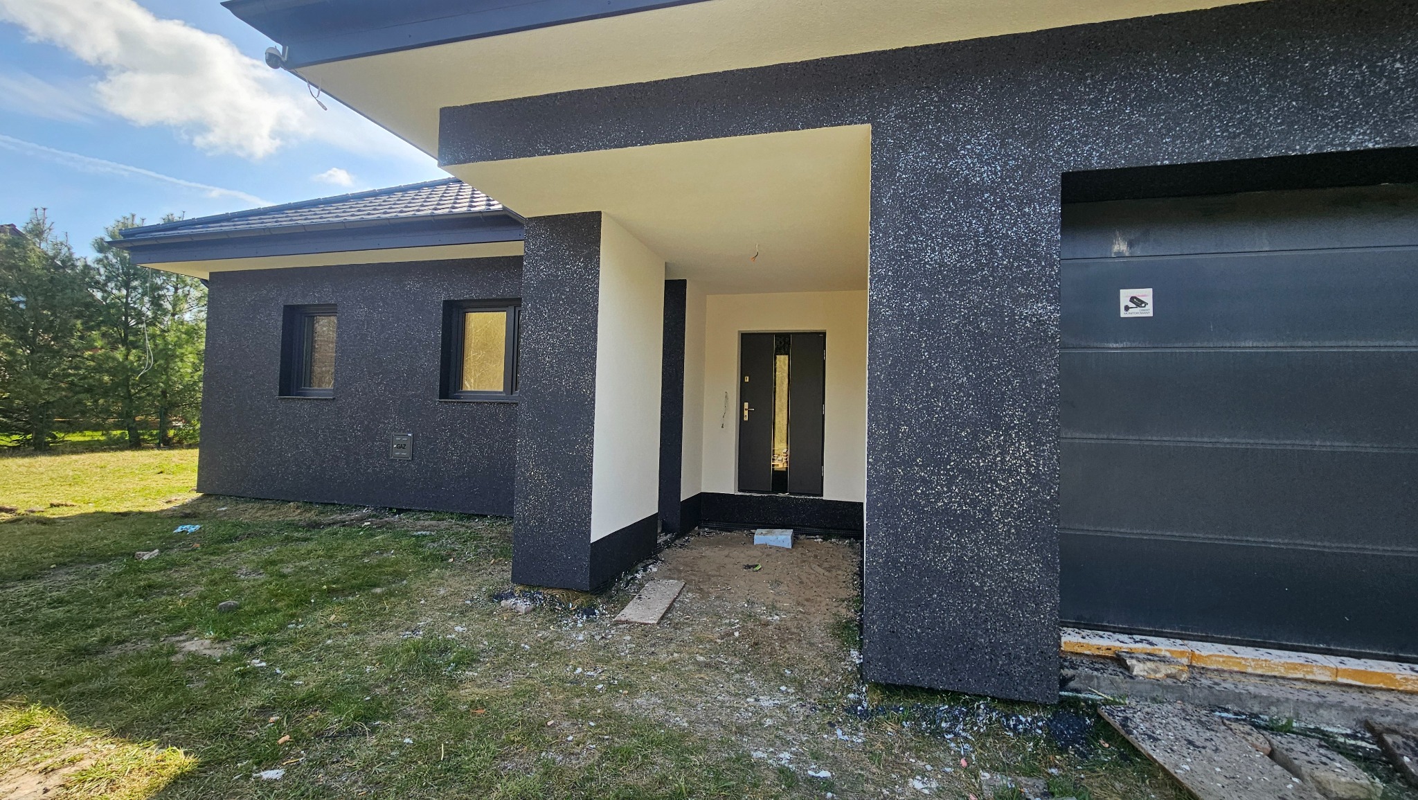 Dom jednorodzinny we Wrocławiu z elewacją w odcieniach szarości i czerni. Widoczny fragment ogrodu i brama garażowa. Charakterystyczna faktura tynku.