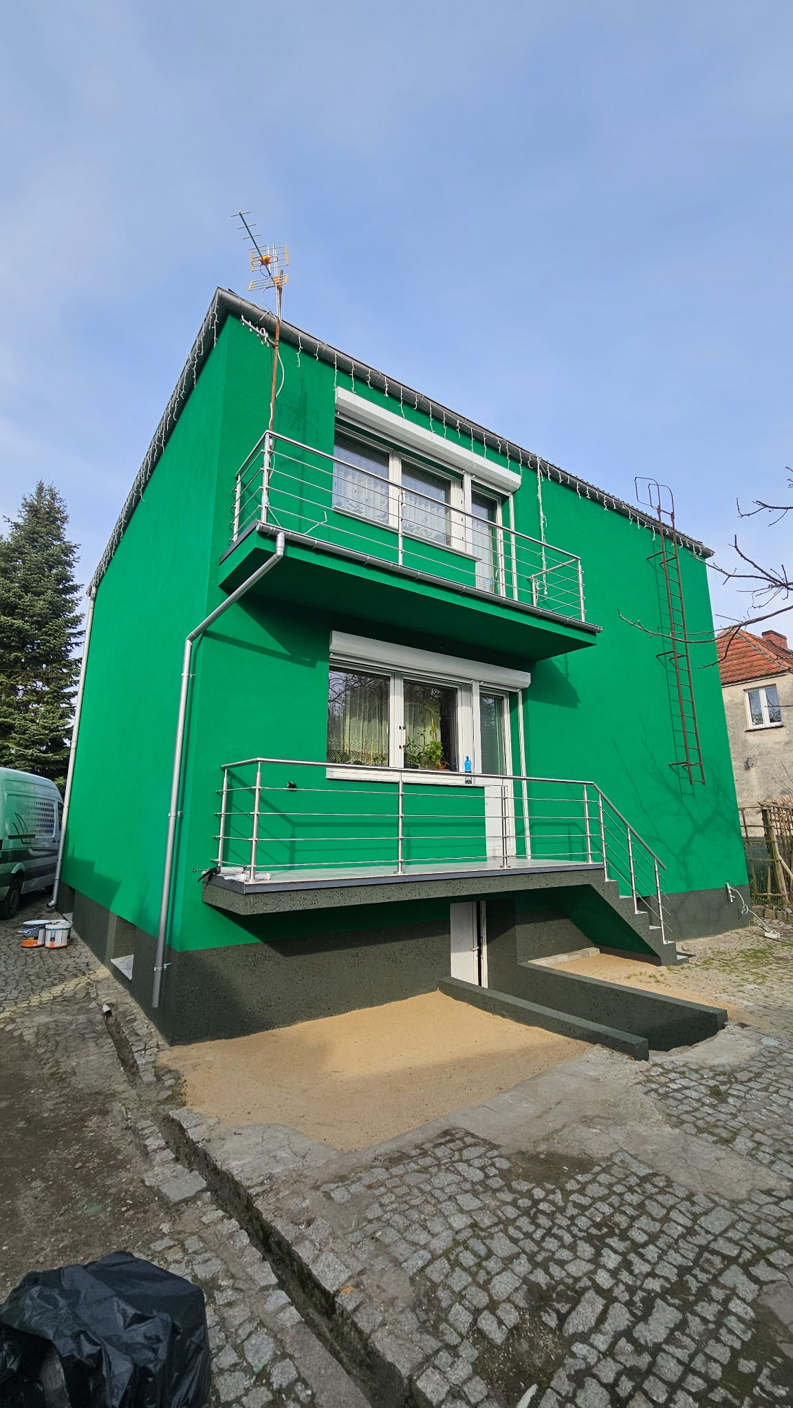 Jaskrawo zielony budynek z balkonami i nierdzewną balustradą, po renowacji elewacji. Widoczne detale wykończenia i drabina na ścianie. Ujęcie z dołu.