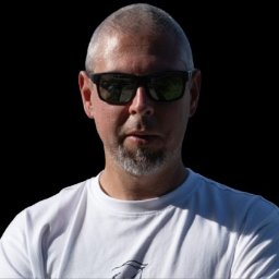 WebAzymut - Portret mężczyzny z krótkimi włosami i brodą, w okularach przeciwsłonecznych i białej koszulce, na czarnym tle. Ujęcie od przodu, ostre światło.