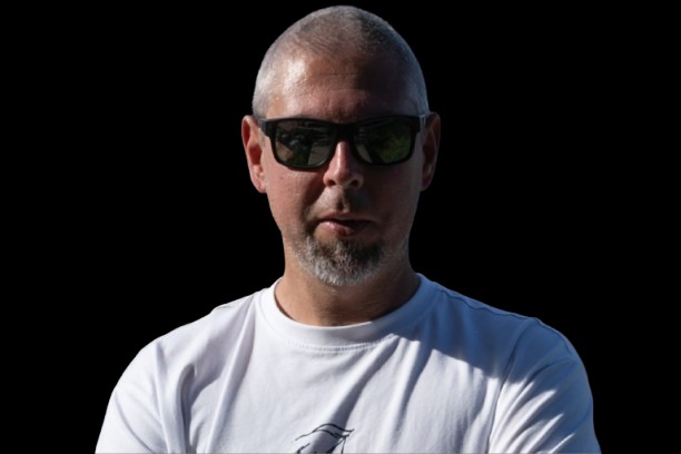 Portret mężczyzny z krótkimi włosami i brodą, w okularach przeciwsłonecznych i białej koszulce, na czarnym tle. Ujęcie od przodu, ostre światło.