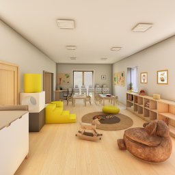 Aleksandra Nycz - Jasne wnętrze sali zabaw z drewnianymi meblami, zabawkami i dekoracjami ściennymi. Widoczne stoliki, krzesełka, regały i zabawki edukacyjne. Przytulna i bezpieczna przestrzeń.