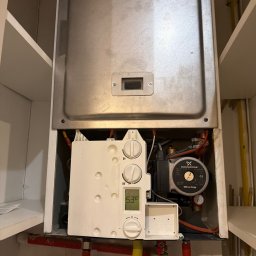 Install-Tech - Wnętrze szafki z zamontowanym piecem gazowym, widoczne rury, pompa obiegowa Grundfos i panel sterowania z wyświetlaczem temperatury.