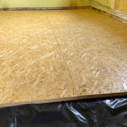 Install-Tech - Ułożona płyta OSB na czarnej folii w pomieszczeniu z żółtymi ścianami. Widoczne kable przy ścianie. Przygotowanie pod podłogę.