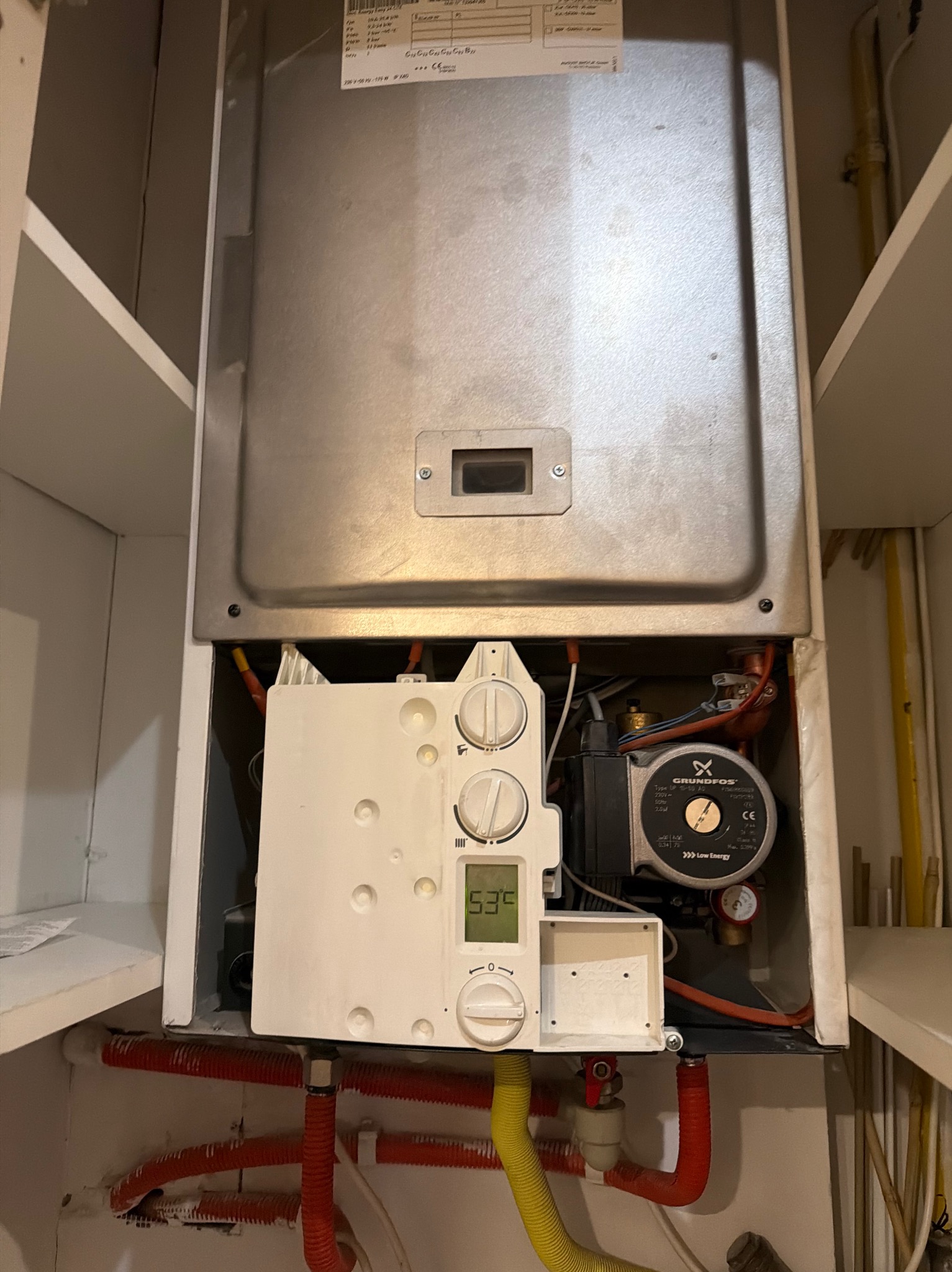 Wnętrze szafki z zamontowanym piecem gazowym, widoczne rury, pompa obiegowa Grundfos i panel sterowania z wyświetlaczem temperatury.