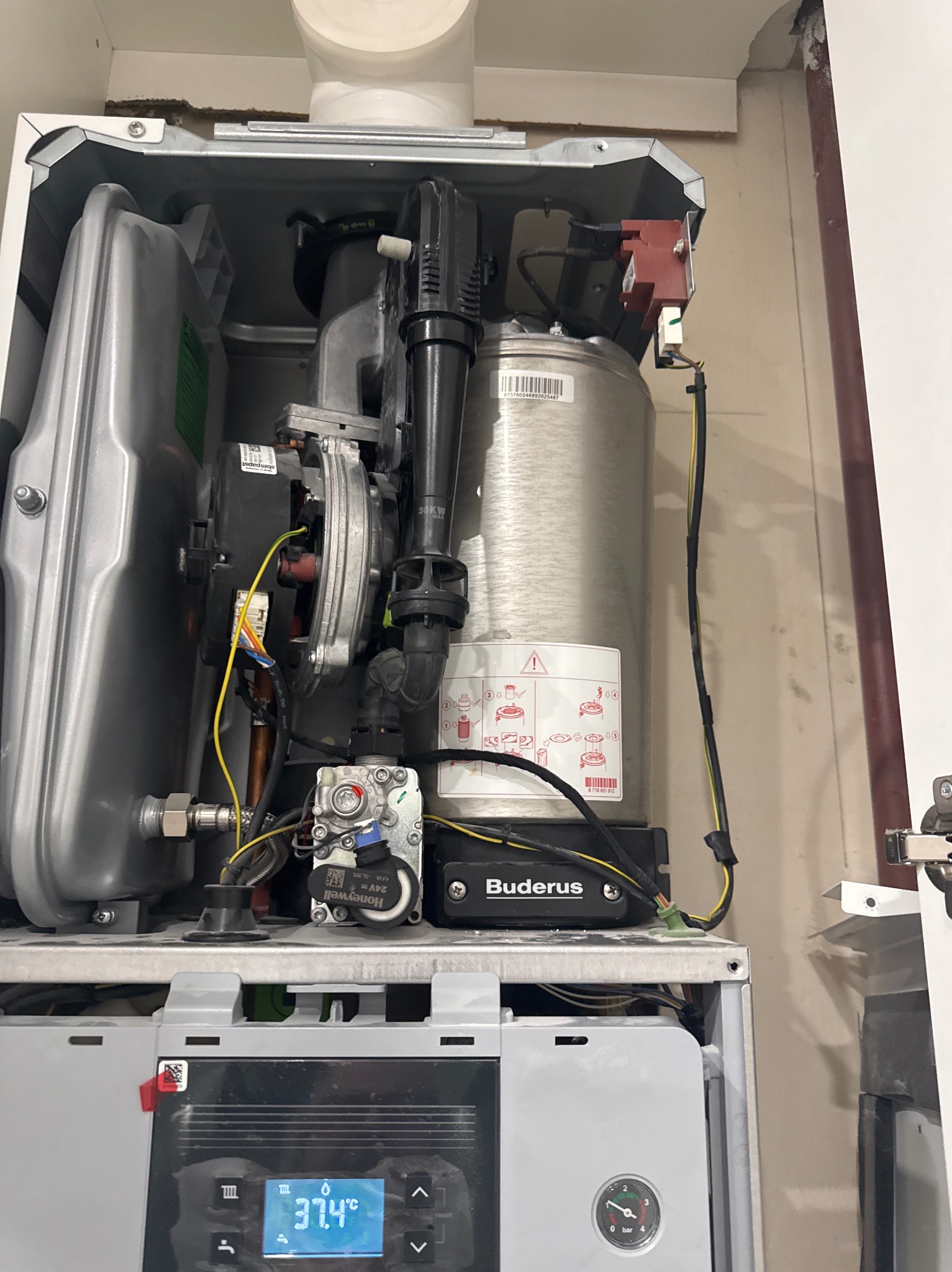 Otwarty piec gazowy marki Buderus z widocznymi elementami instalacji, przewodami i wyświetlaczem wskazującym temperaturę 37.4°C. Widoczne komponenty ebmpapst i Honeywell.