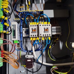 ELEKTROMECHANIKA Dominik Sobczyński - Rozdzielnica elektryczna w szarej obudowie z widocznymi bezpiecznikami, przewodami oznaczonymi żółtymi końcówkami i elementami sterującymi. Widoczne napisy 'ODBIORY'.