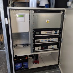 ELEKTROMECHANIKA Dominik Sobczyński - Otwarta szafa sterownicza z akumulatorem, bezpiecznikami i modułami kontrolnymi. Widoczna kamera Dahua na górze. Elementy instalacji elektrycznej wewnątrz obudowy.