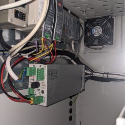 ELEKTROMECHANIKA Dominik Sobczyński - Wnętrze szafy sterowniczej z modułami zasilania, przewodami i wentylatorem. Widoczne złącza, kable i elementy elektroniczne. Detal instalacji elektrycznej w obudowie.