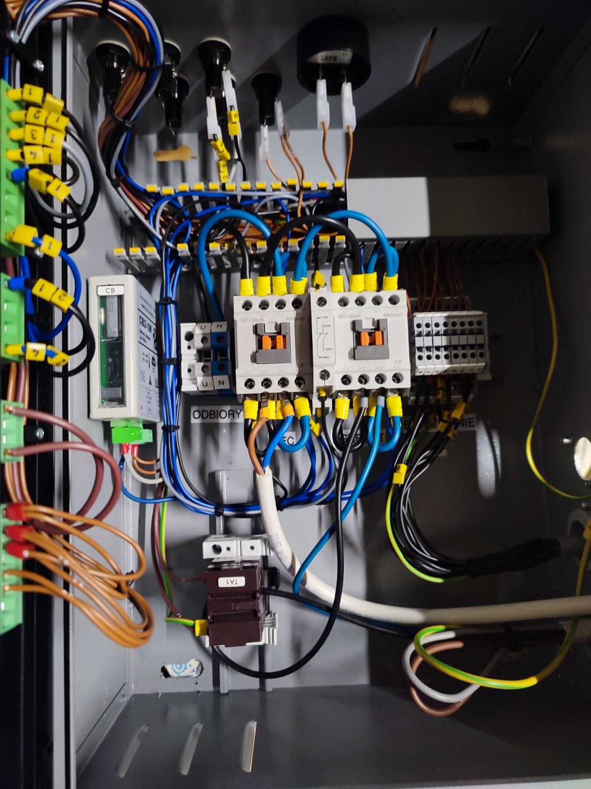 Rozdzielnica elektryczna w szarej obudowie z widocznymi bezpiecznikami, przewodami oznaczonymi żółtymi końcówkami i elementami sterującymi. Widoczne napisy 'ODBIORY'.