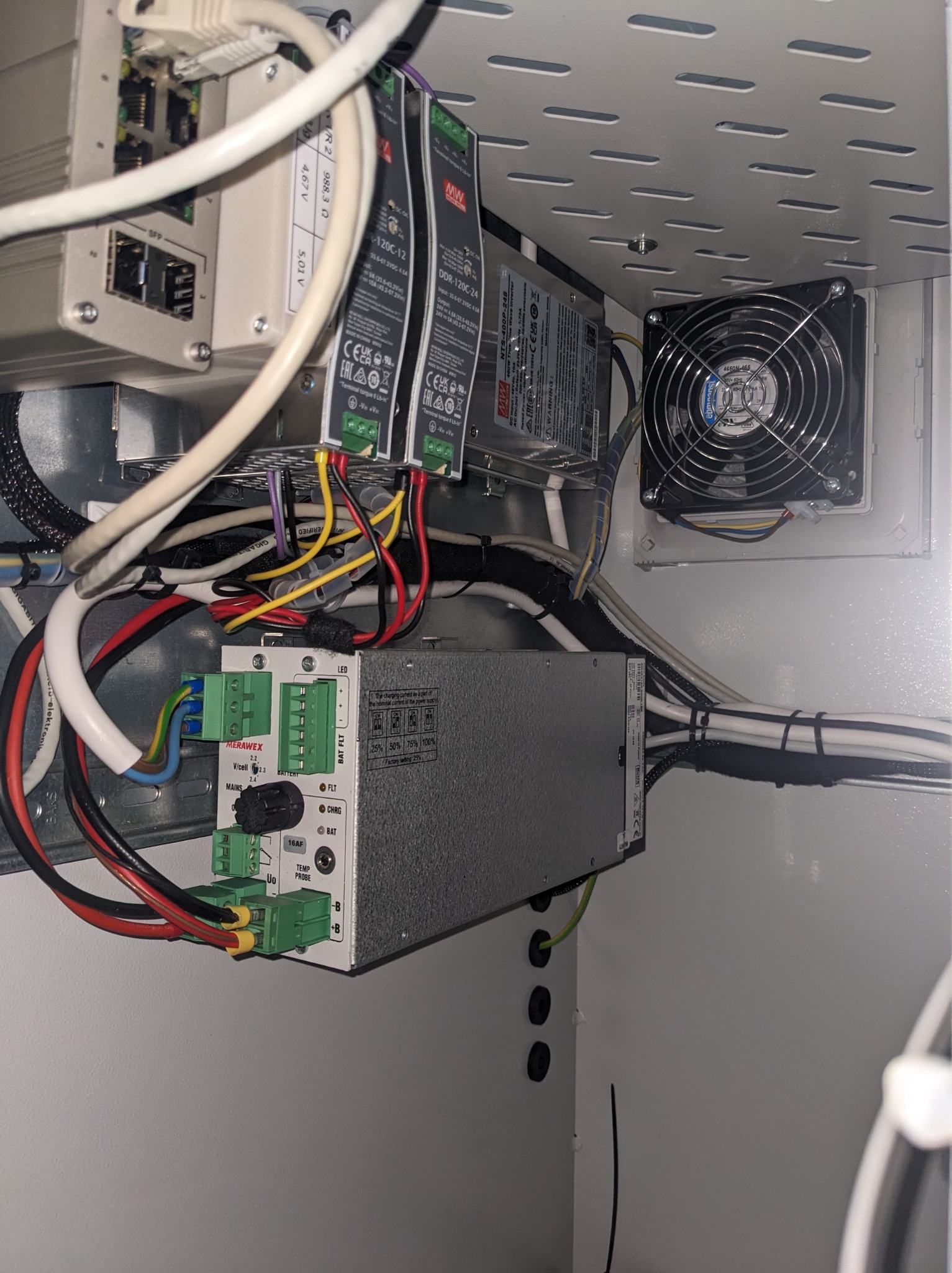 Wnętrze szafy sterowniczej z modułami zasilania, przewodami i wentylatorem. Widoczne złącza, kable i elementy elektroniczne. Detal instalacji elektrycznej w obudowie.