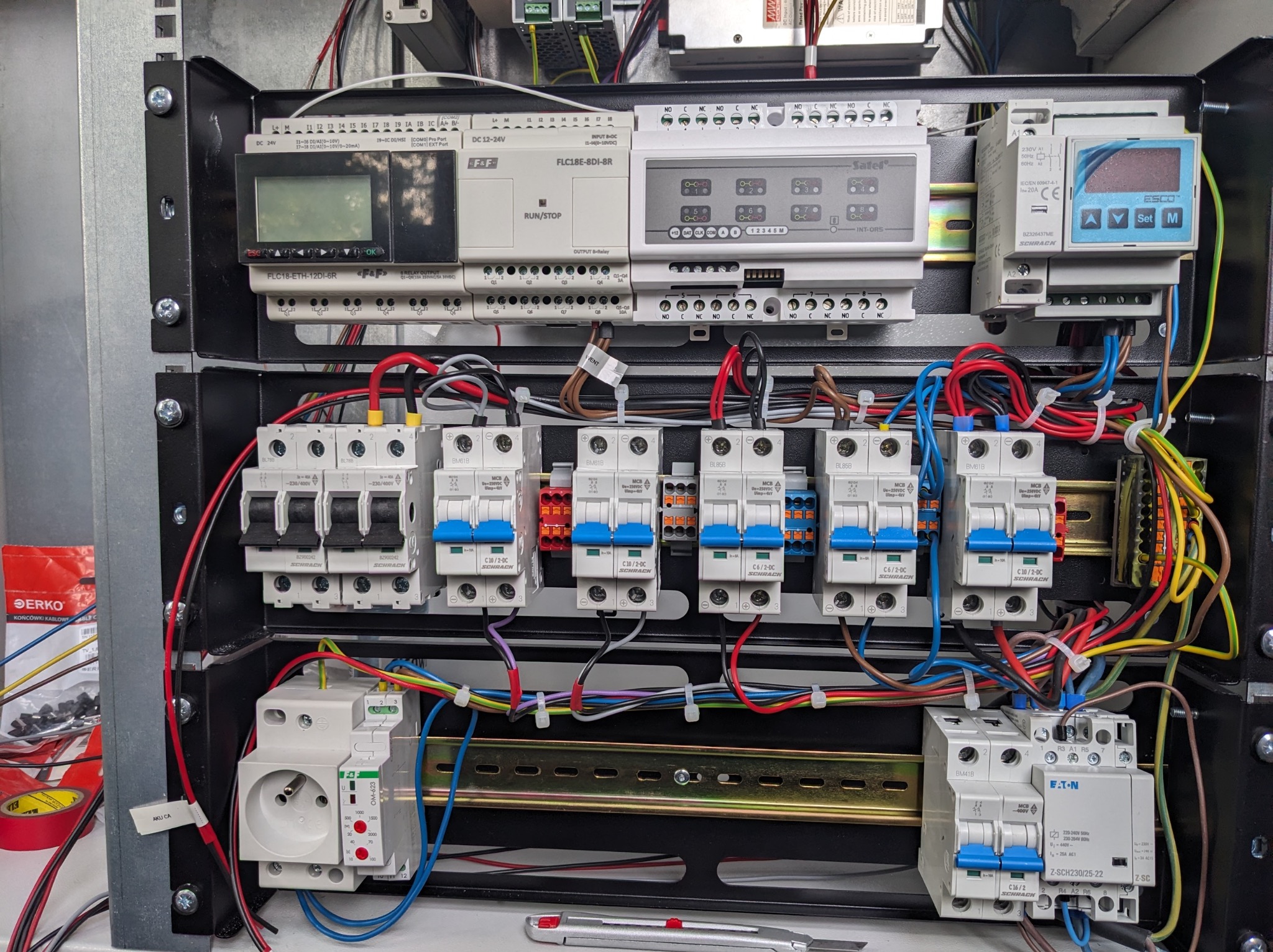 Rozdzielnica elektryczna z zabezpieczeniami, sterownikami i okablowaniem w metalowej obudowie. Widoczne bezpieczniki Schrack, sterowniki F&F i Satel.