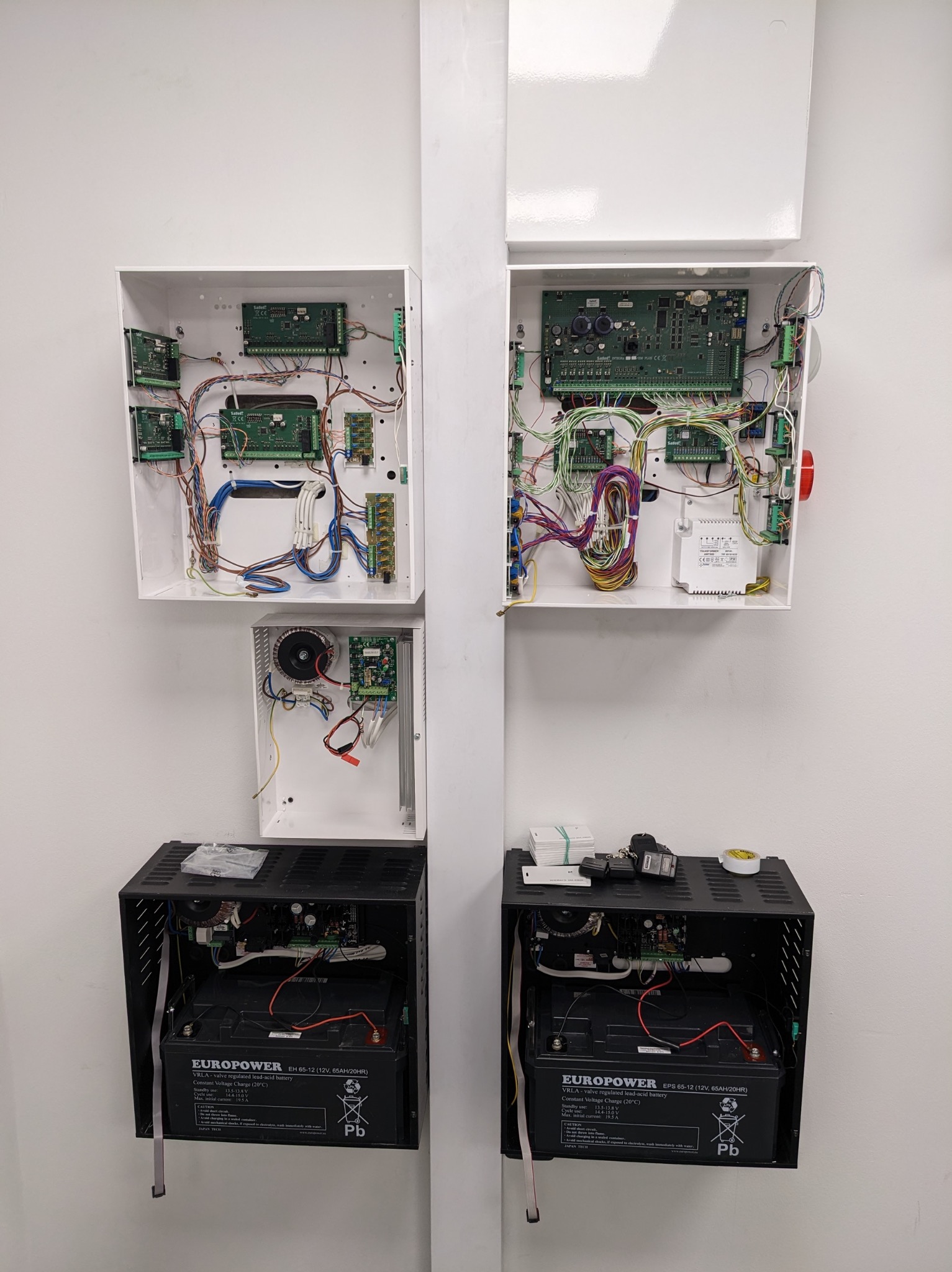 Dwa panele alarmowe z elektroniką i akumulatorami w białych i czarnych obudowach, zamontowane na ścianie. Widoczne kable i podzespoły.