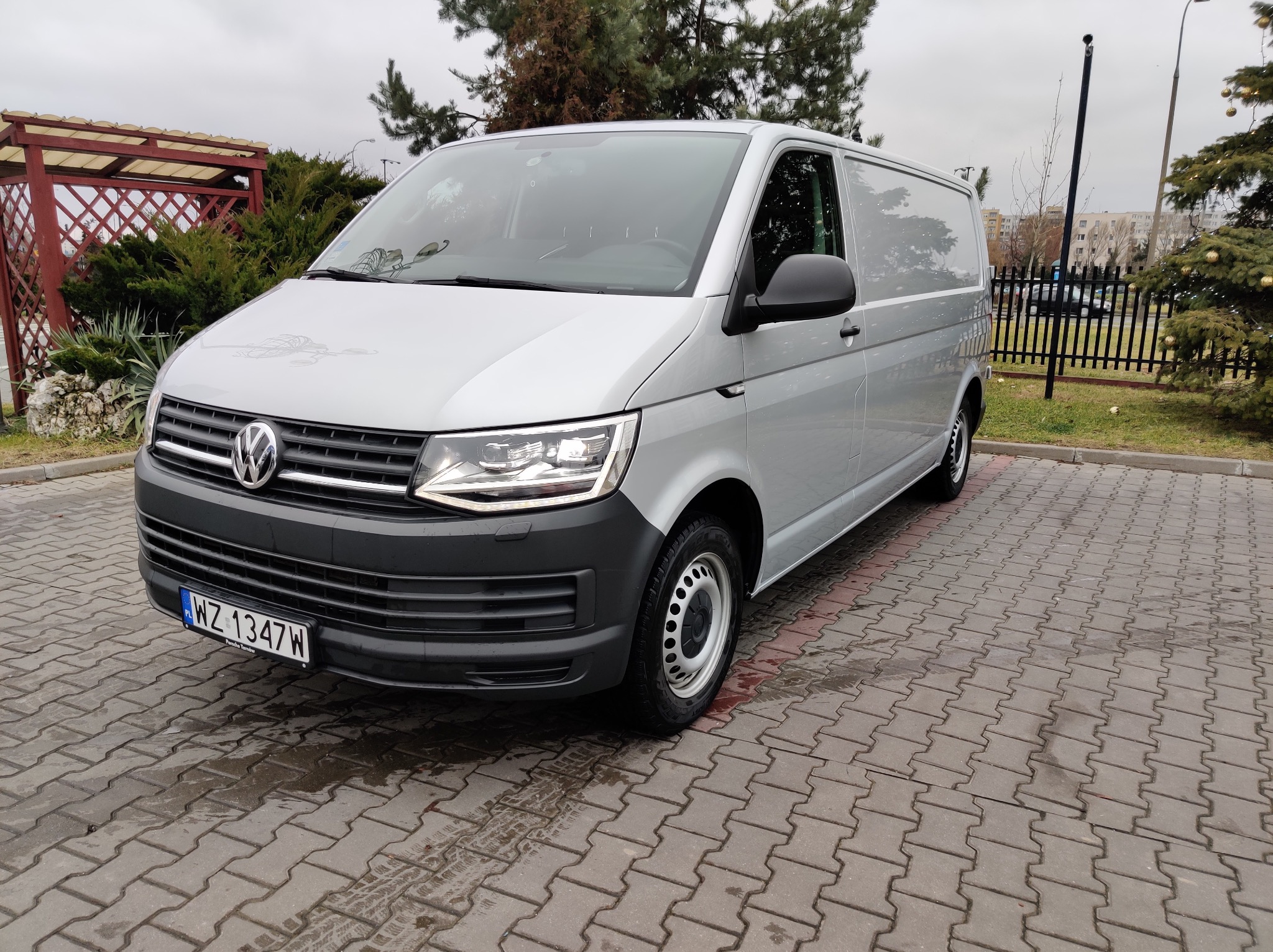Srebrny Volkswagen Transporter T6 na kostce brukowej, z widoczną tablicą rejestracyjną i elementami otoczenia, jak drzewa i budynki w tle. Pochmurny dzień.
